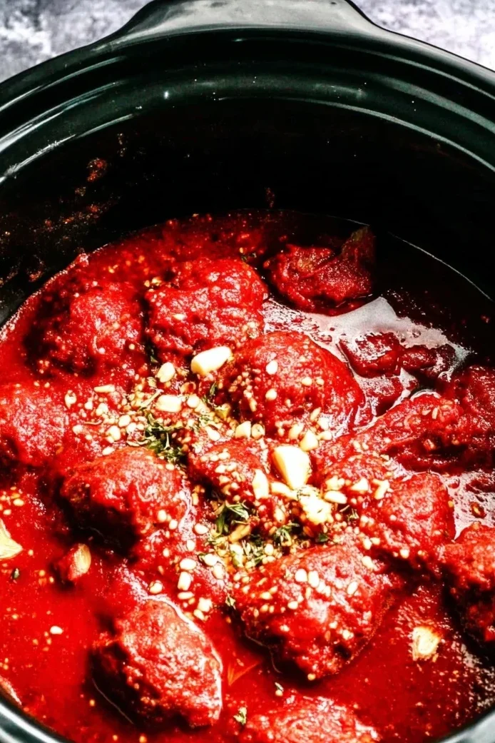 Cooked meatballs simmering in rich marinara sauce inside a black slow cooker, topped with herbs and garlic